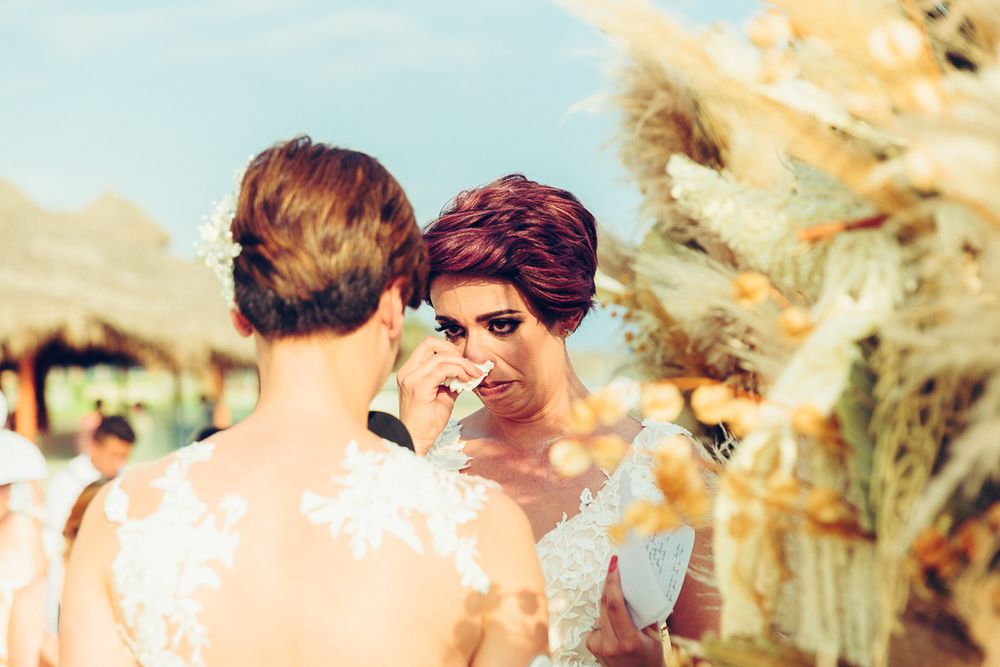 Two people in white lace dresses share an emotional moment at an outdoor wedding venue.