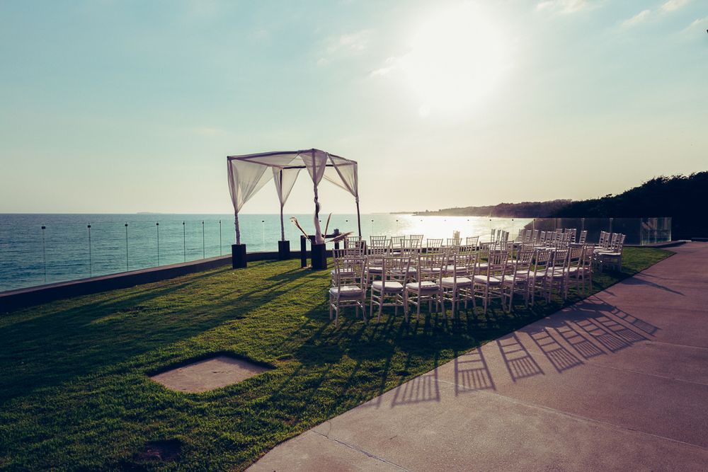 Outdoor beach wedding ceremony setup with white chairs and canopy arch against ocean backdrop at sunset.