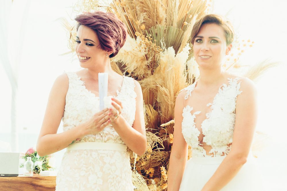 Two people in elegant white lace wedding dresses stand beside decorative dried pampas grass.