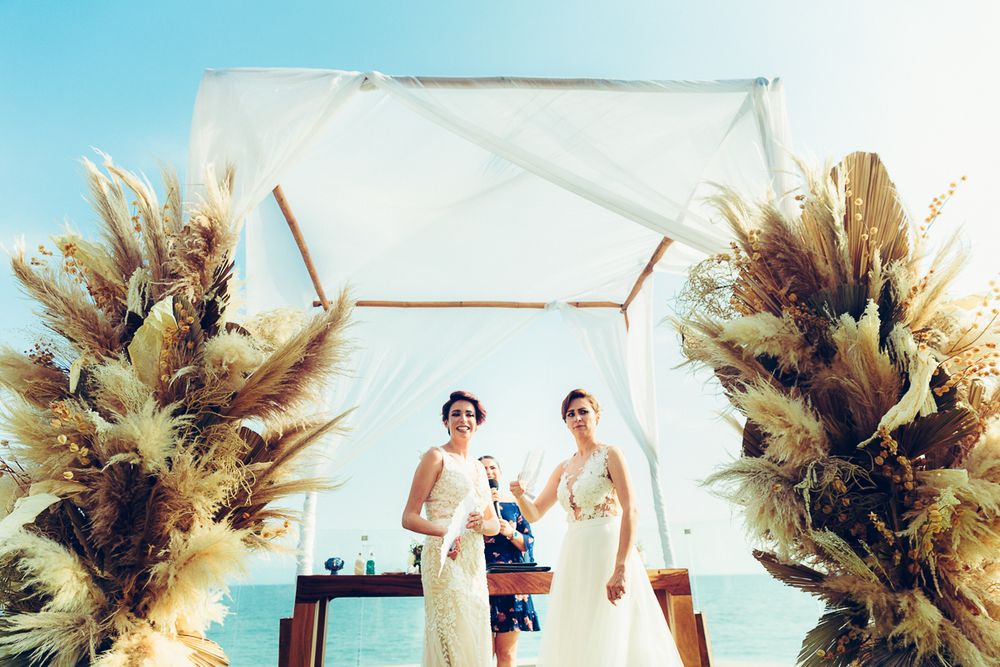A seaside wedding ceremony setup with dramatic pampas grass arrangements framing a white arch.