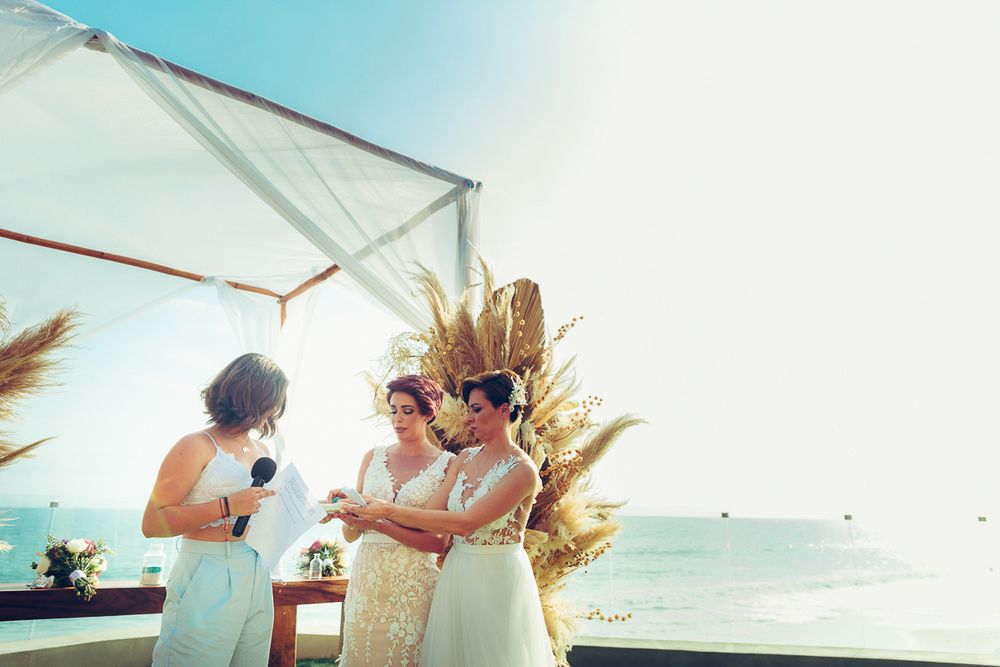 A beachside wedding ceremony with attendees gathered under a white canopy with dried floral decor.