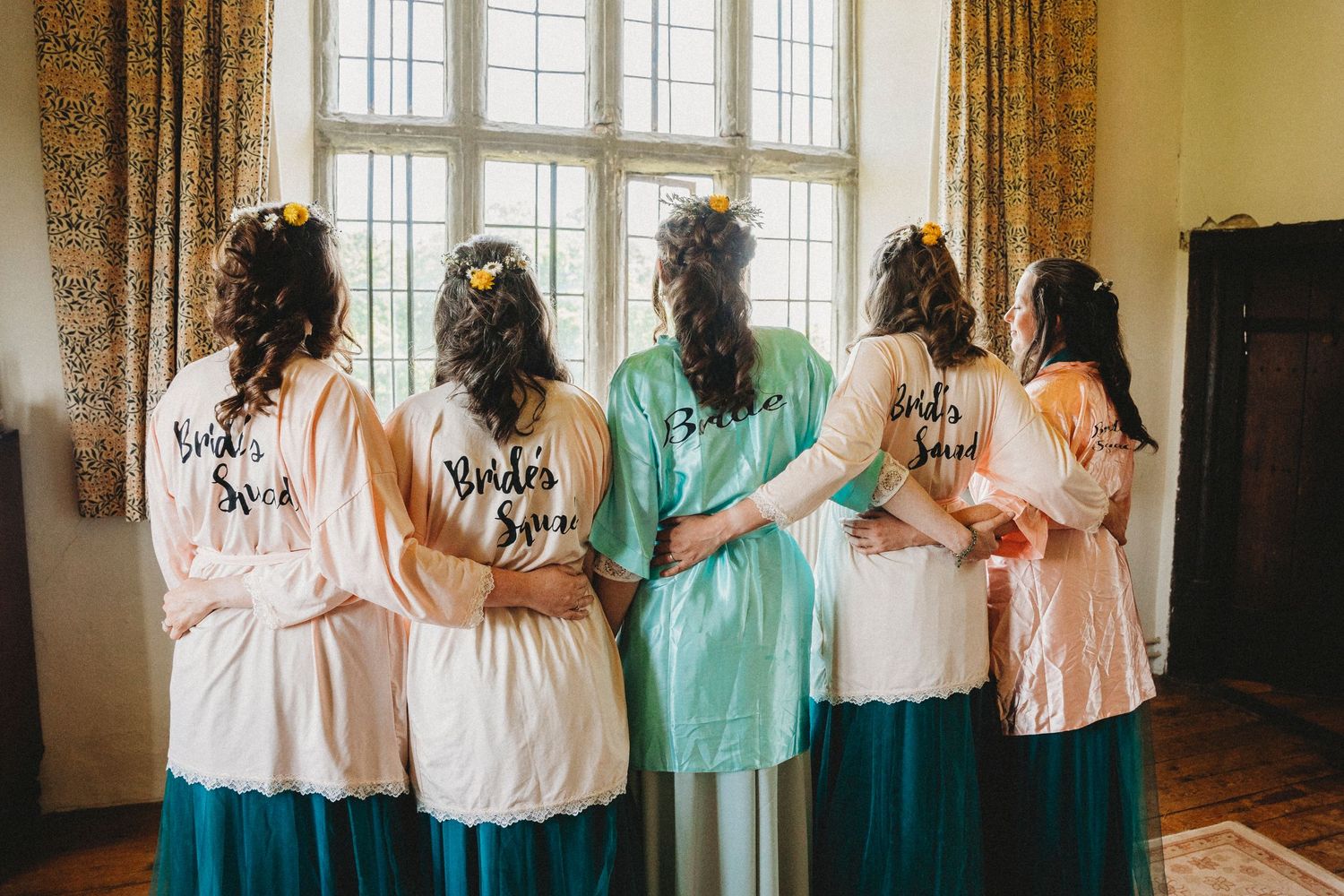 Group in pastel dresses standing by large windows with floral curtains.