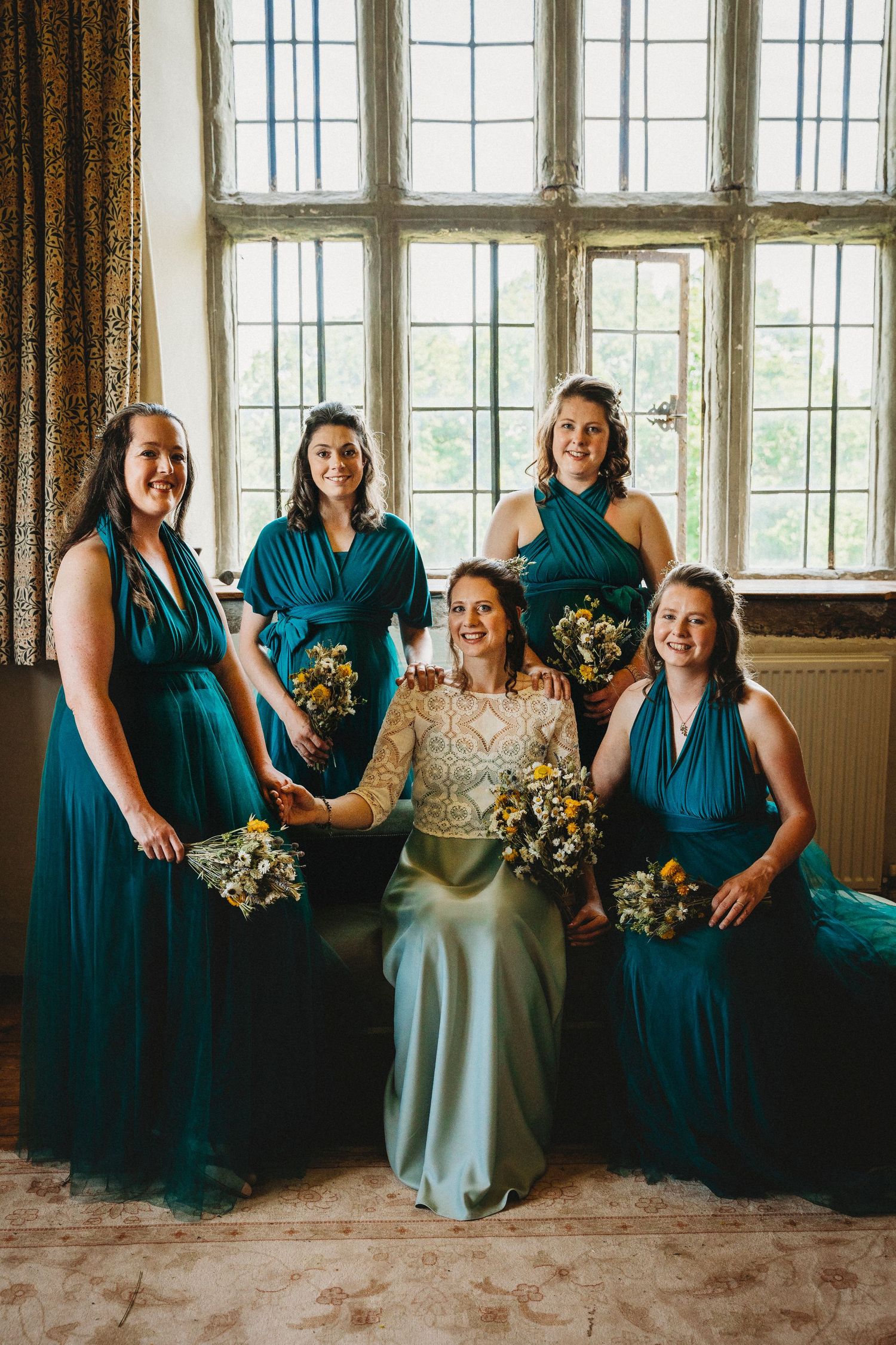 Wedding party wearing teal bridesmaid dresses and white gown pose by vintage windows in historic venue.