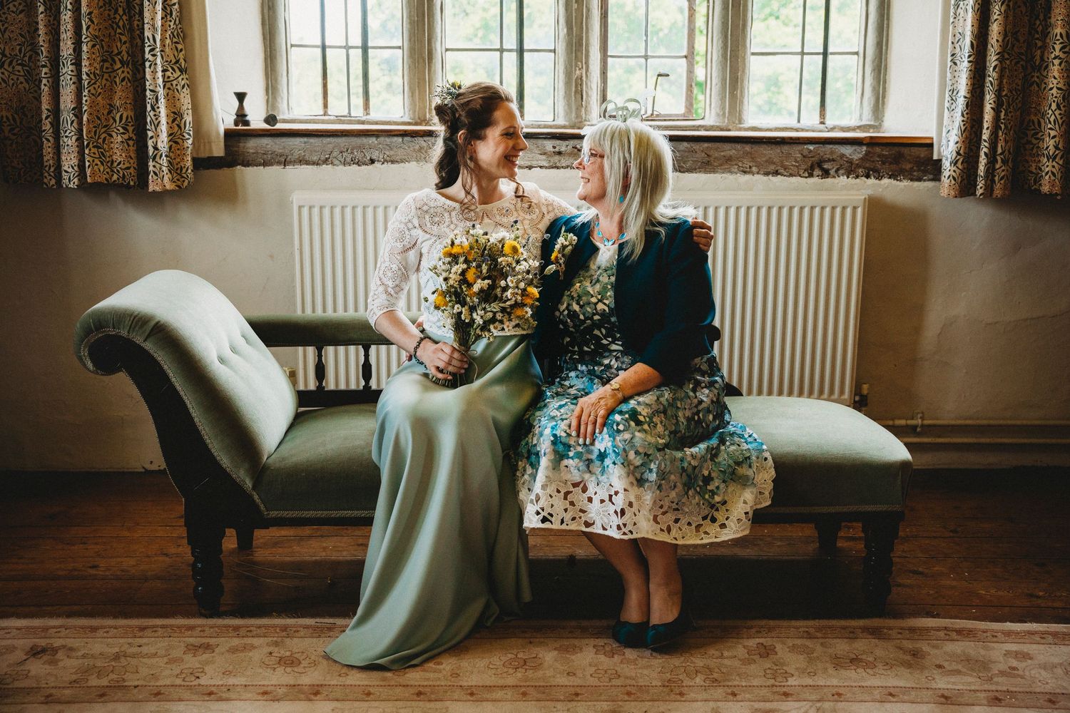 Wedding party relaxes on antique furniture by large windows in rustic indoor setting.