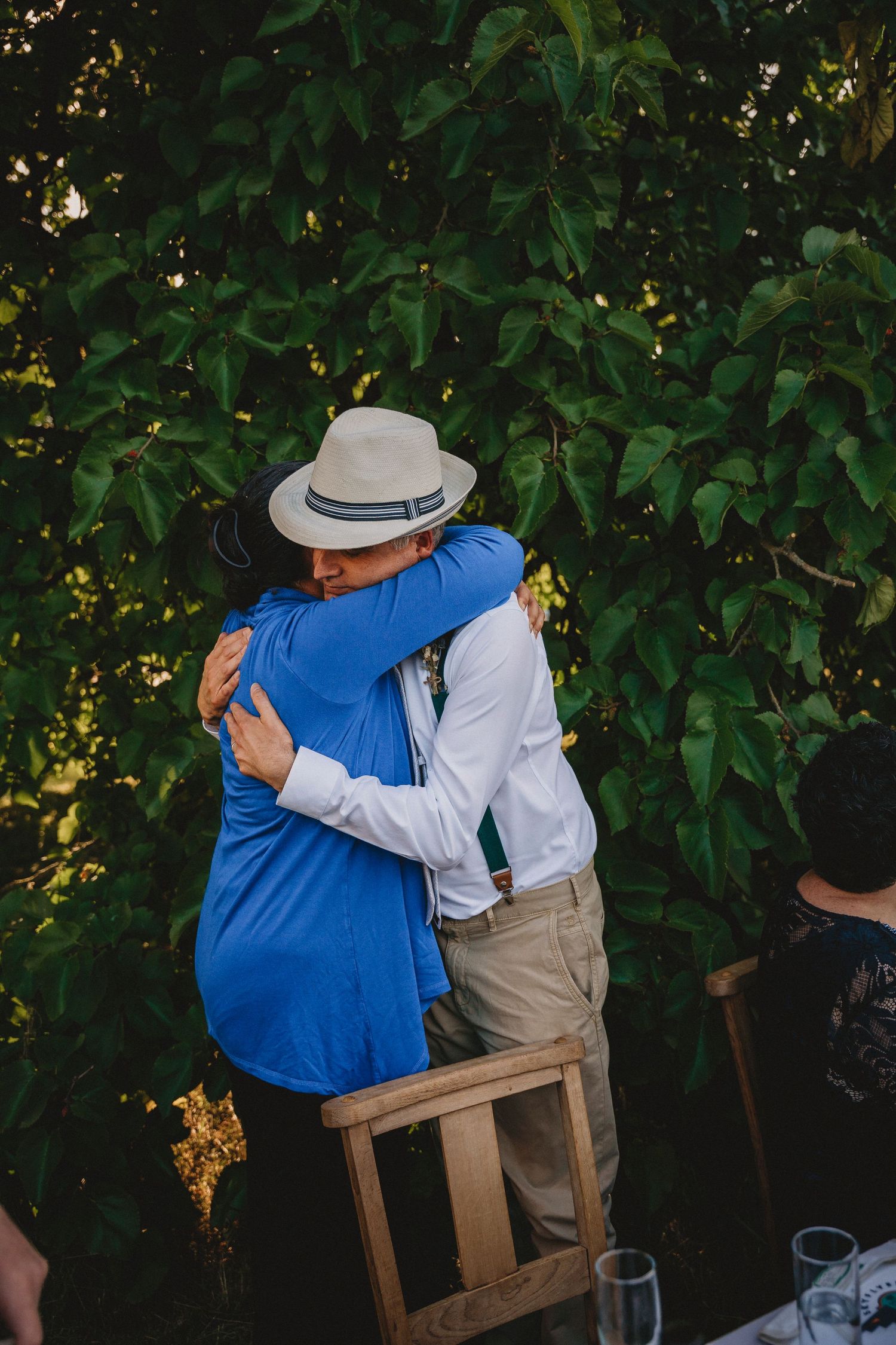 Two people sharing an intimate embrace while seated on wooden chairs at an outdoor event.