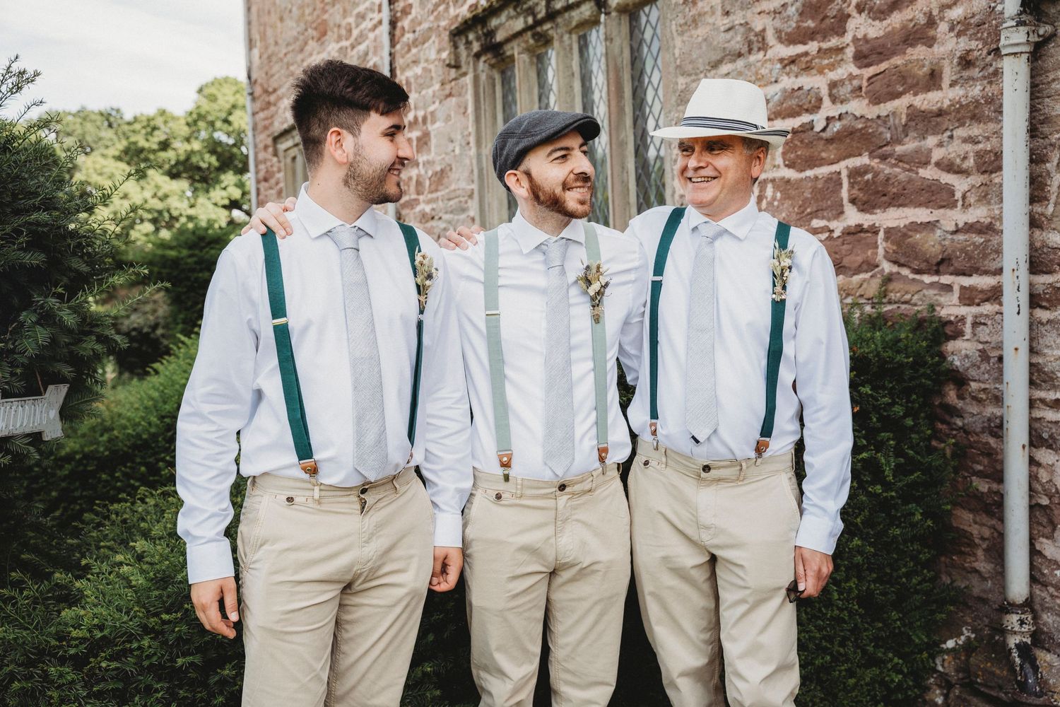 Groomsmen in suspenders and khaki pants pose together outside historic building.