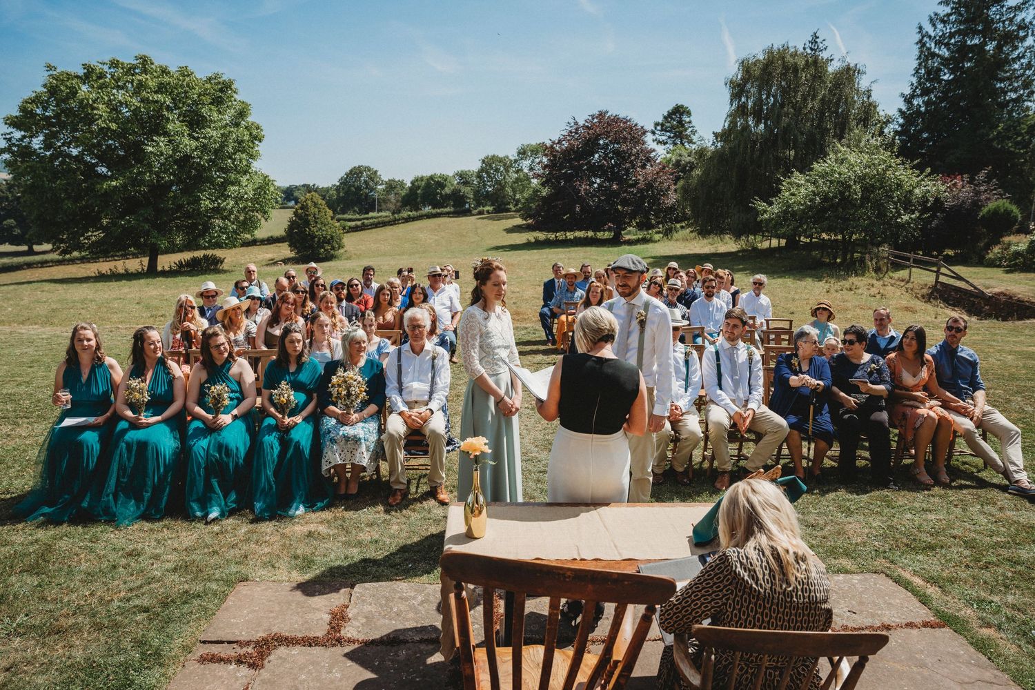 Wedding ceremony guests seated on lawn with castle and gardens in background.