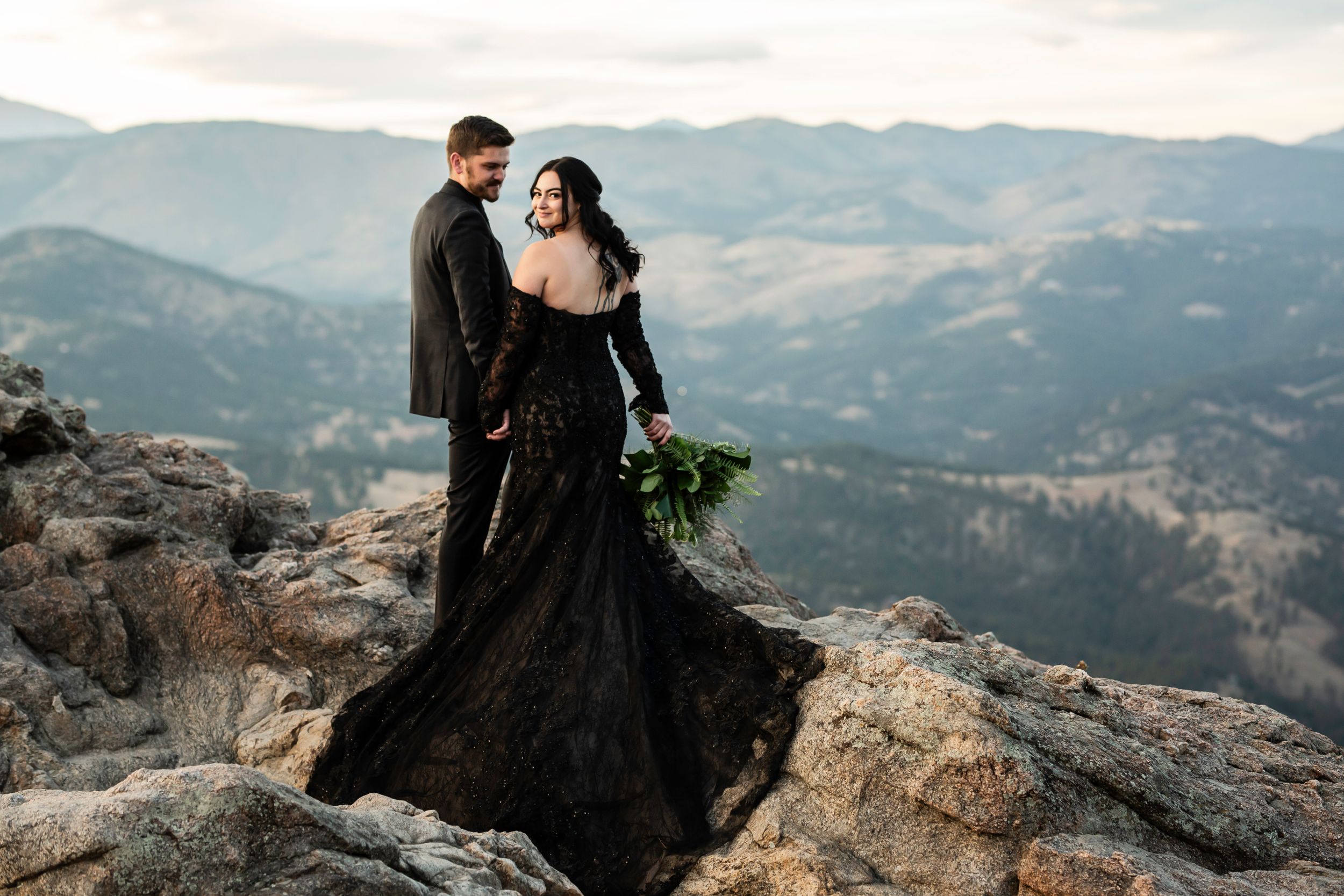 A couple in dramatic black attire poses on rocky mountain overlook during their wedding shoot.