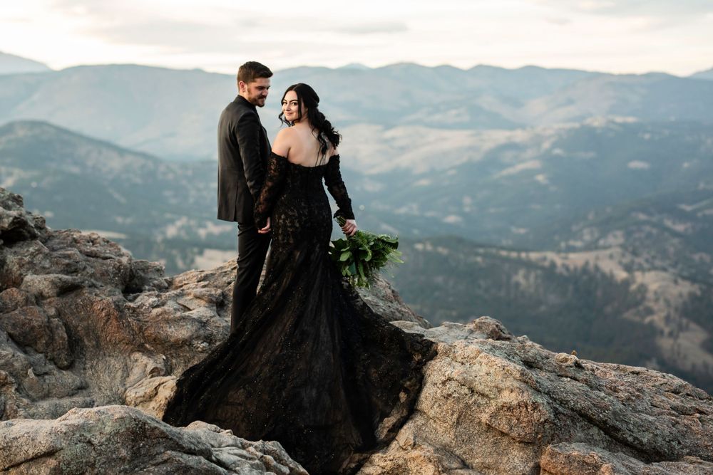 A couple in dramatic black attire poses on rocky mountain overlook during their wedding shoot.