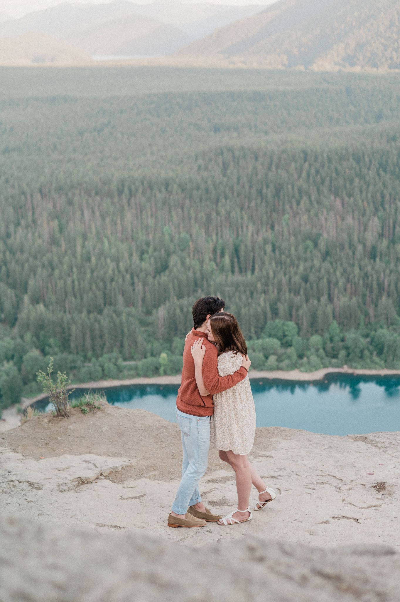 A couple shares a romantic moment on a cliff overlooking a serene mountain lake surrounded by evergreen forest.