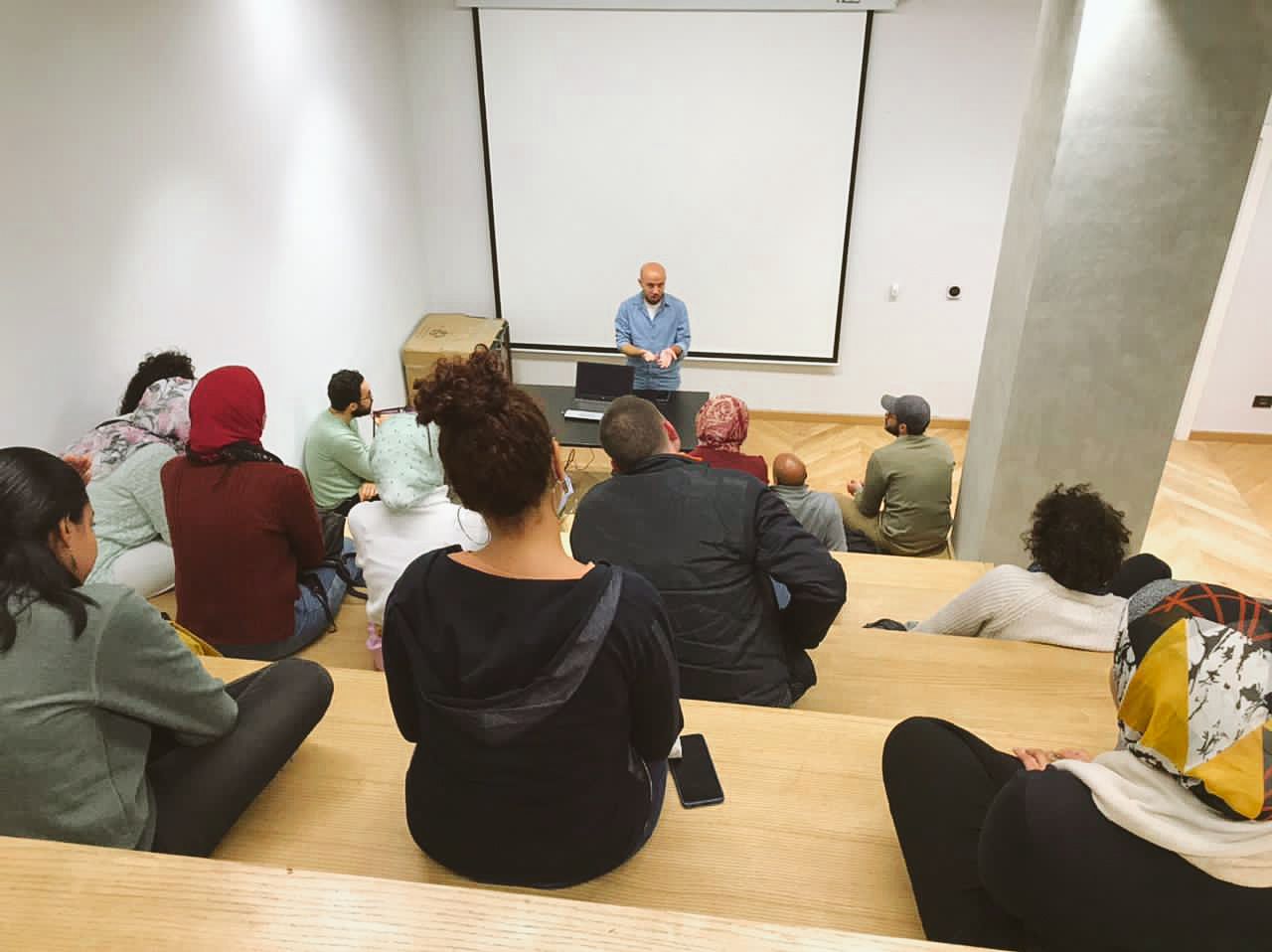 A group of students sit on tiered seating while attending a lecture in a classroom with a projection screen.