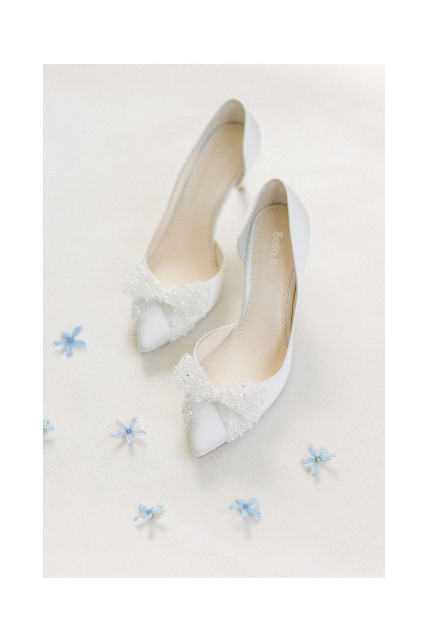 White bridal flats with delicate lace details and bows displayed with small blue flowers.