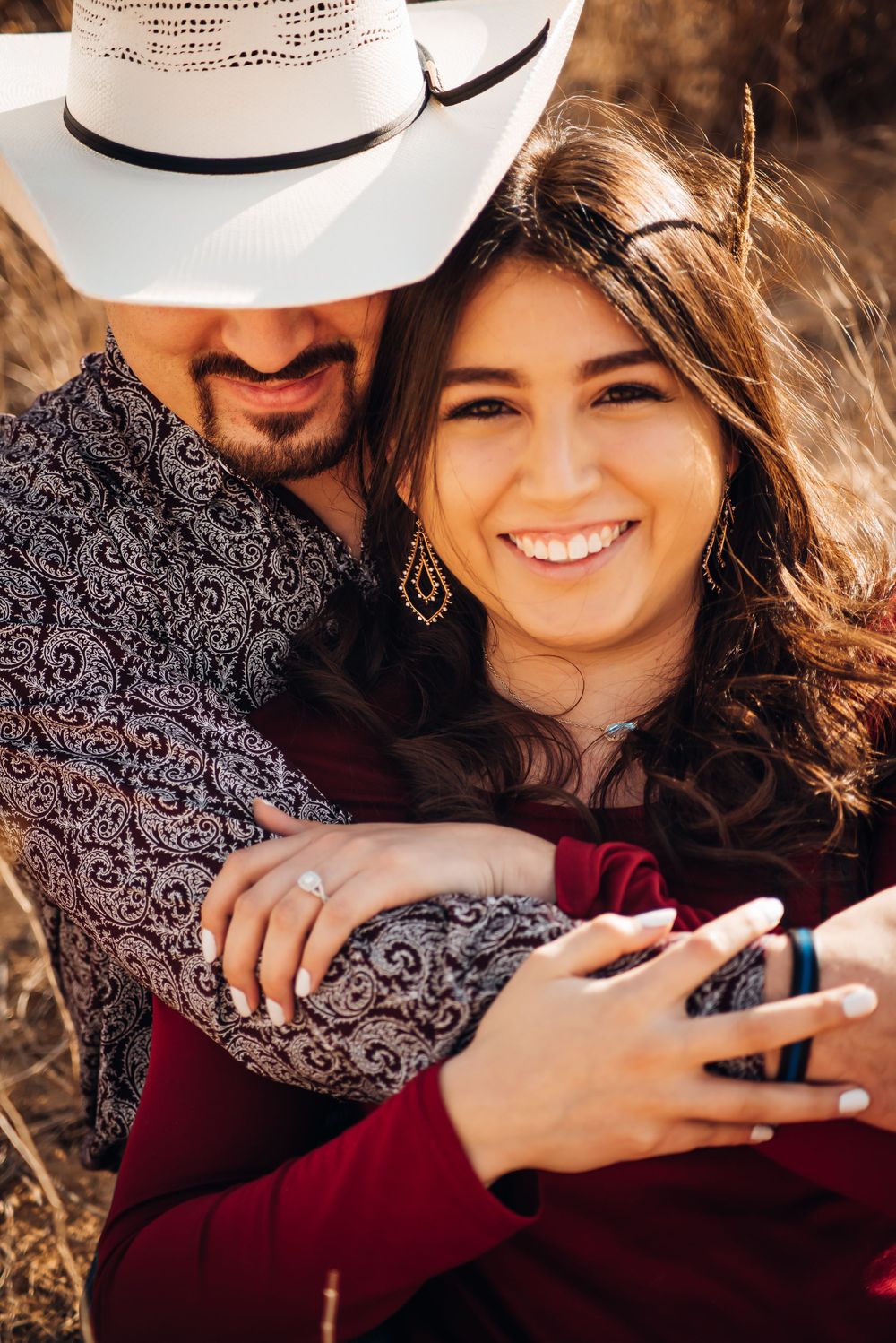 Romantic couple embraces in a field at sunset, wearing western attire with a white cowboy hat and burgundy dress.