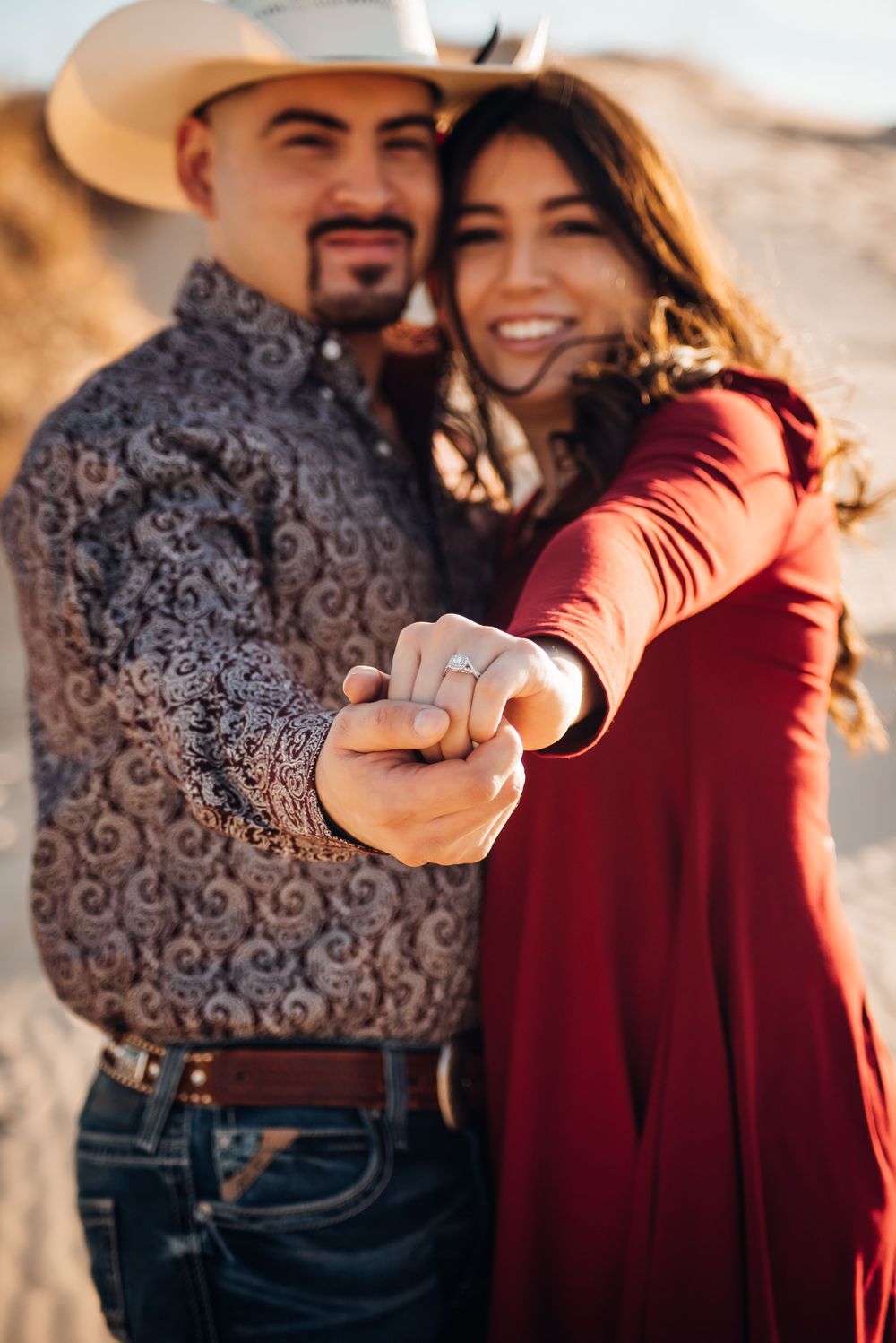 Couple in western wear smiles at camera while holding hands in desert setting.
