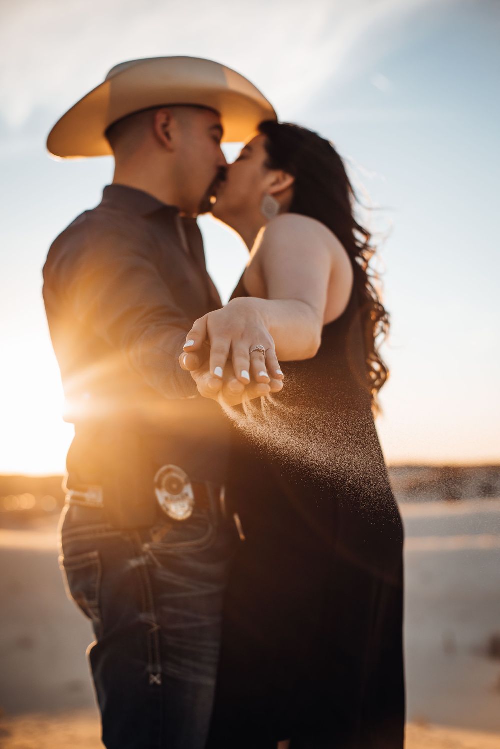 Silhouetted couple embraces during golden hour with cowboy hat visible against bright sky.