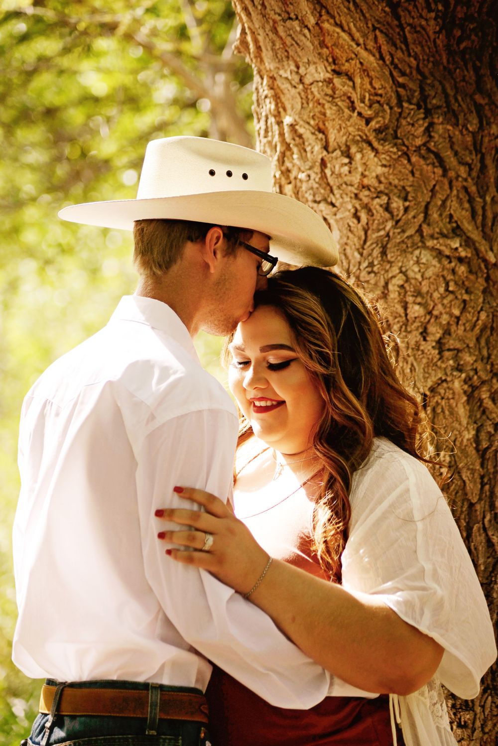 A romantic couple embraces under a tree in a sunlit outdoor setting.