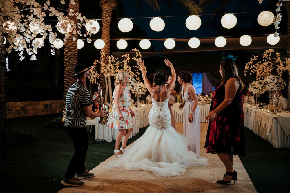 Wedding party dances at outdoor reception under glowing lanterns and palm trees.