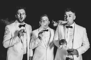 Three men in white tuxedos striking playful poses with drinks at a formal event.