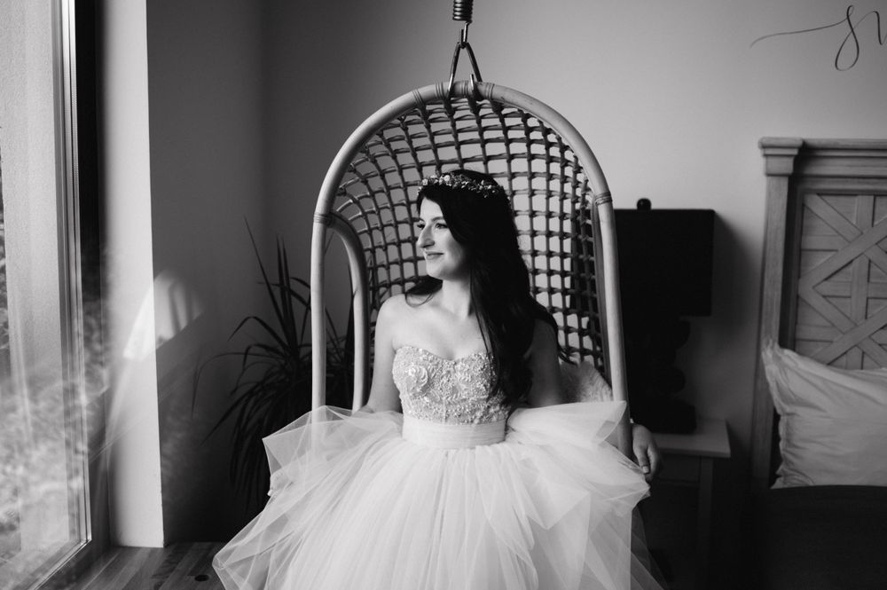Black and white portrait of a bride in a hanging rattan chair beside large windows.