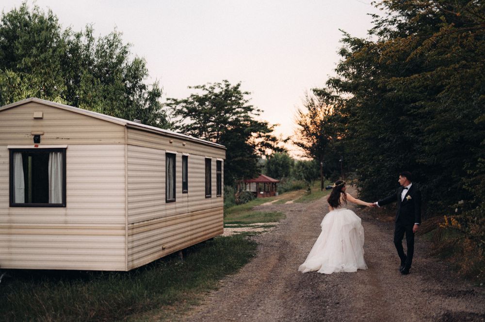 Wedding couple walking on dirt path past mobile home during sunset.