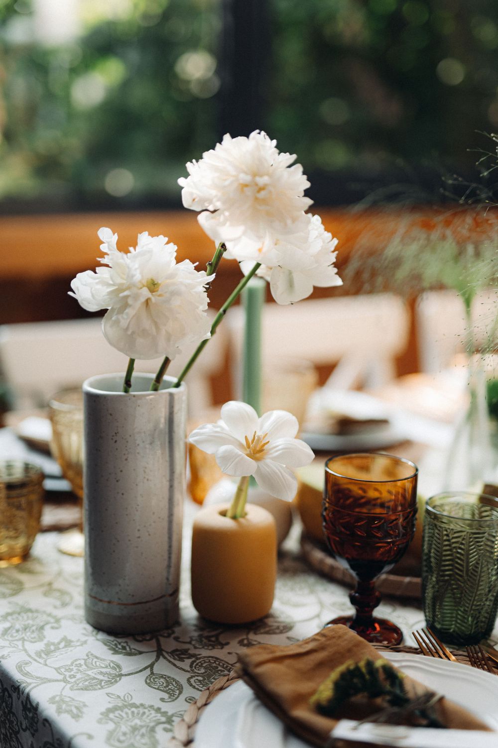 Elegant table setting with white peonies in a gray vase, colored glassware, and candlelight.