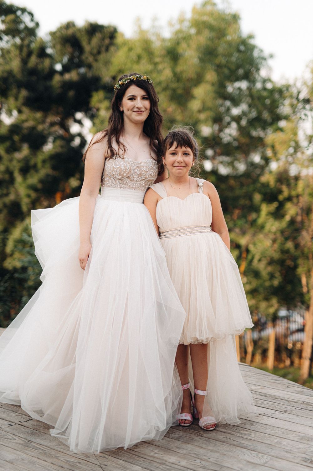 Two figures in flowing white dresses pose together on a wooden deck during a wedding celebration.