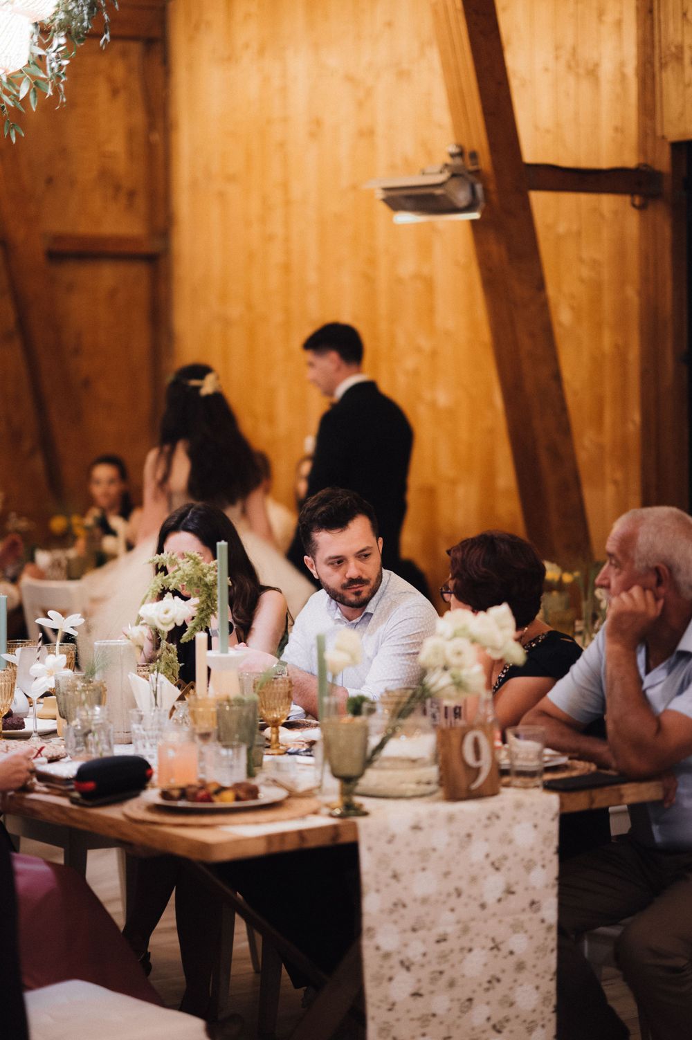 Wedding guests enjoy dinner and conversation at rustic wooden tables inside a barn-style reception venue.
