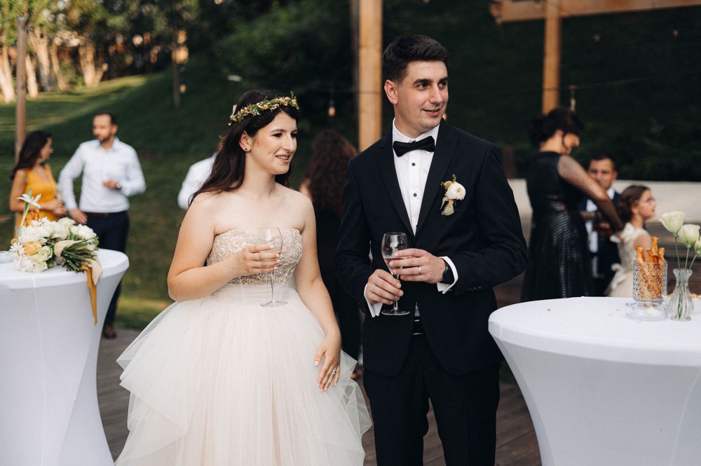 A newlywed couple in formal attire holding champagne glasses during their outdoor wedding reception.
