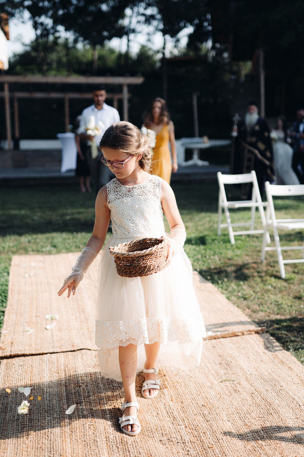 A young flower girl in a white dress scatters petals down an outdoor aisle runner.