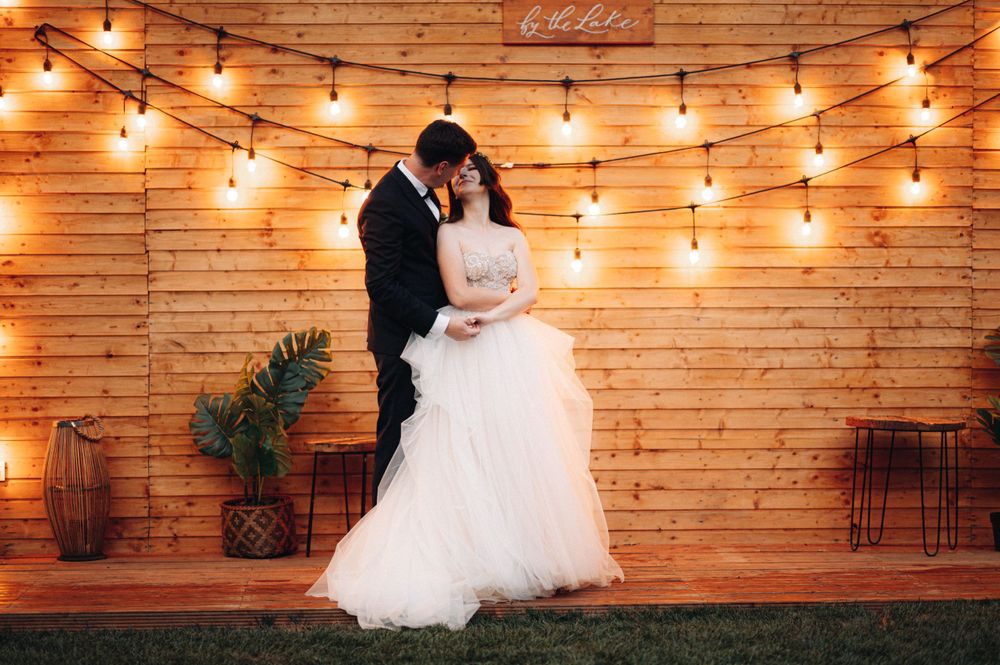Couple embraces under warm string lights against a wooden wall backdrop at their wedding reception.