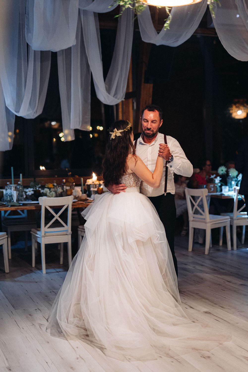 A couple dances in an elegantly decorated reception hall with flowing white drapes and ambient lighting.