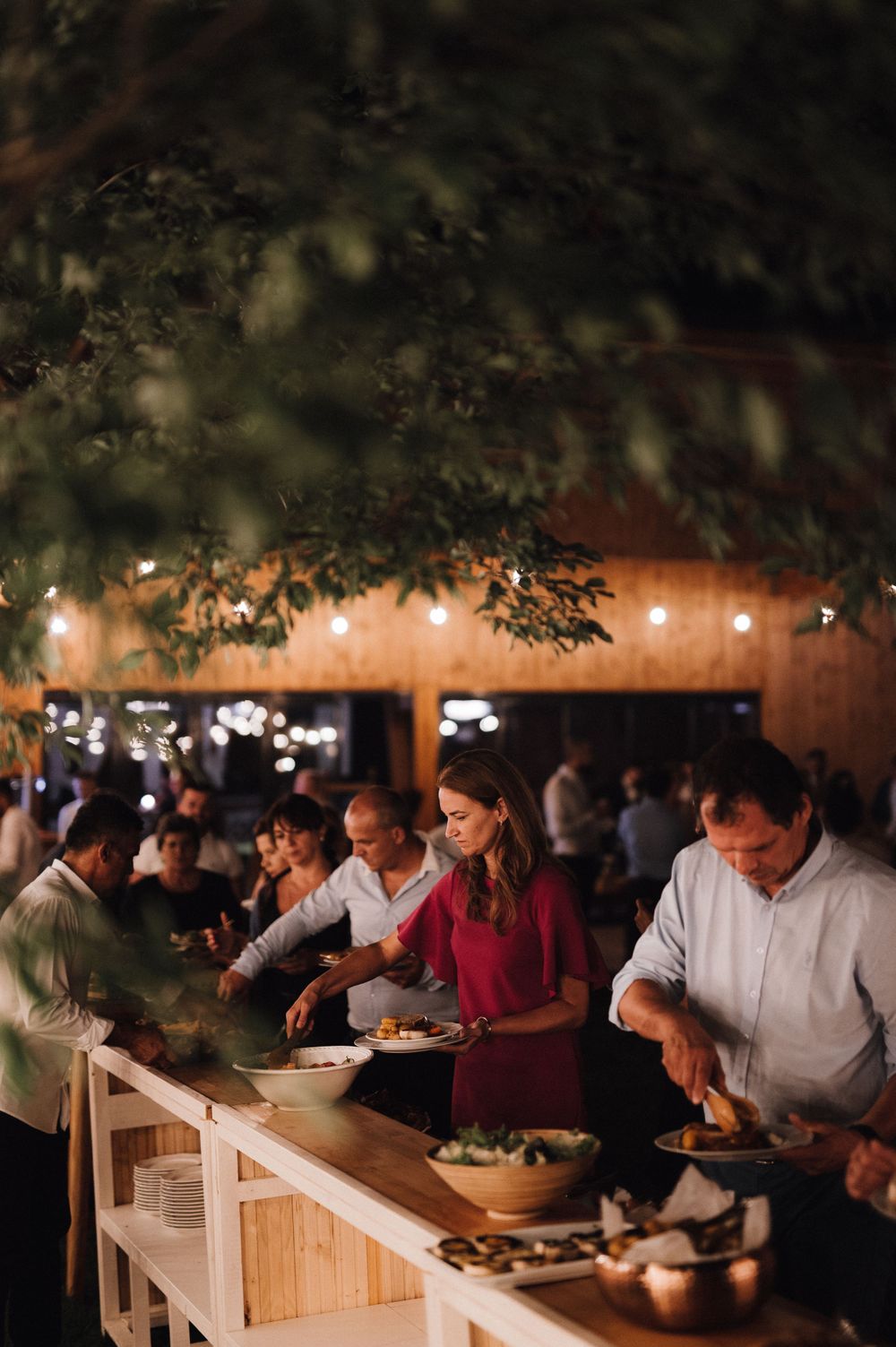 Wedding guests serving themselves at a buffet line under string lights and wooden decor.
