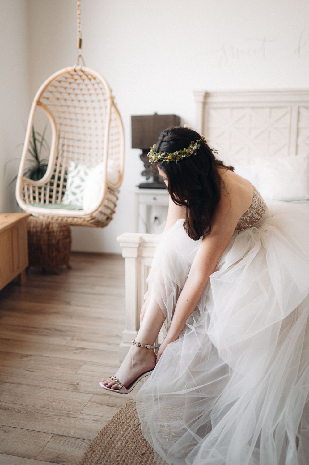 Bride in a strapless wedding gown putting on shoes near a hanging rattan chair.