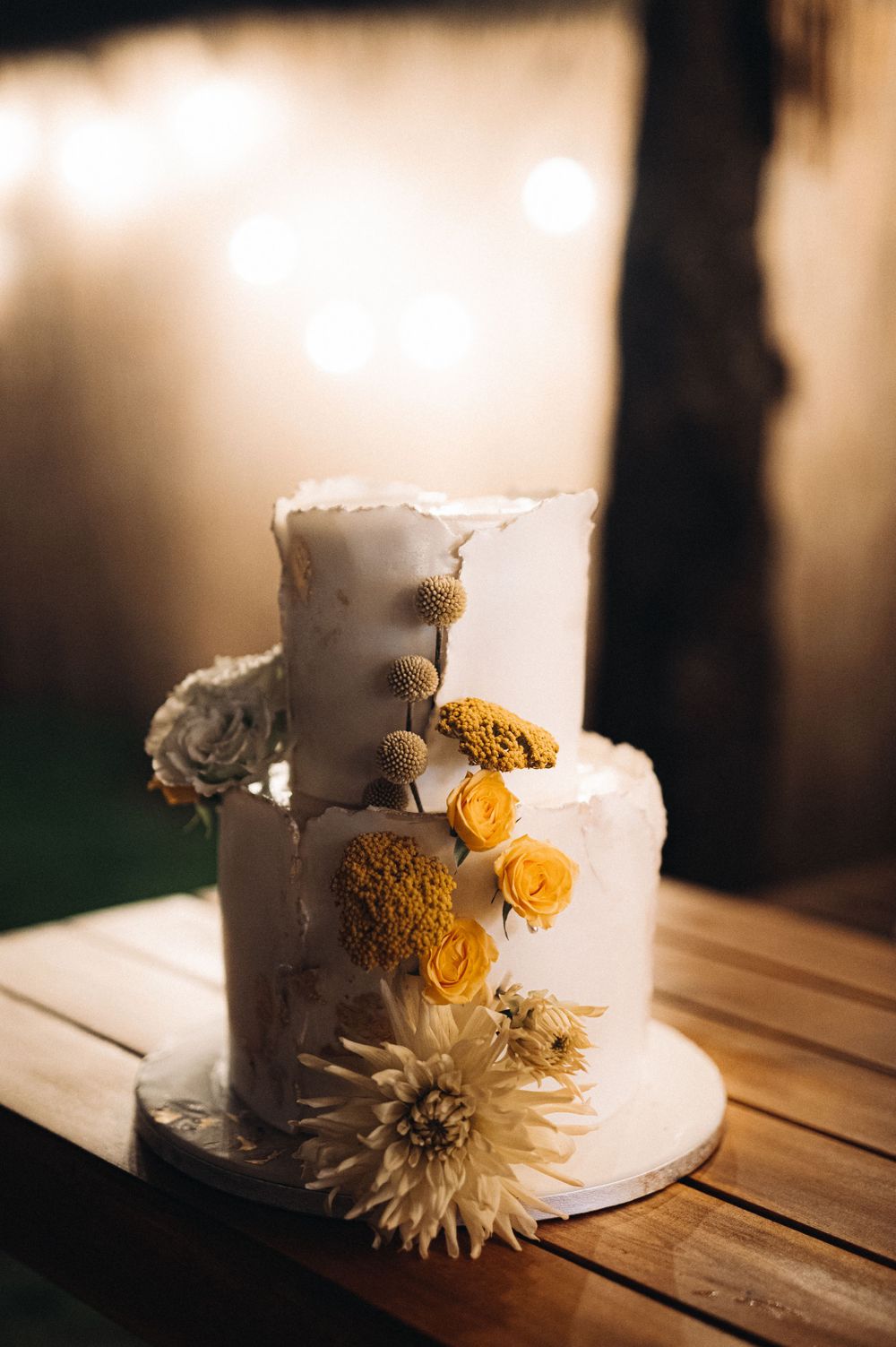 Two-tiered white wedding cake decorated with yellow roses and a large dahlia on wooden table.