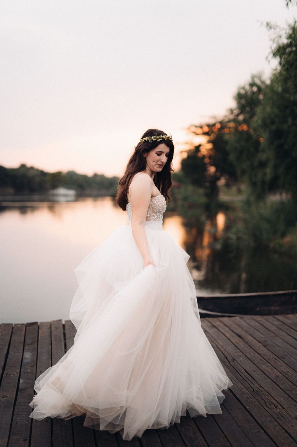 An elegant bride in a flowing white tulle gown poses on a lakeside dock at dusk.