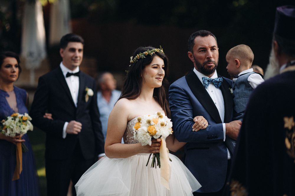 A couple walking during an elegant outdoor wedding ceremony with a bride in a white ball gown holding yellow flowers.
