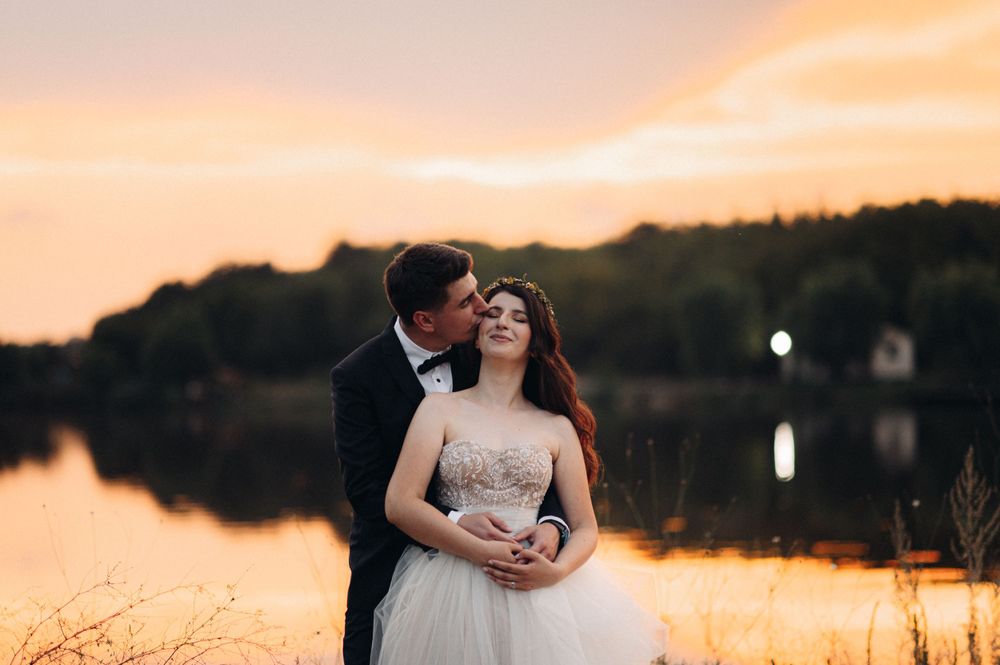 Romantic sunset portrait of newlyweds by a serene lake with golden sky reflections.