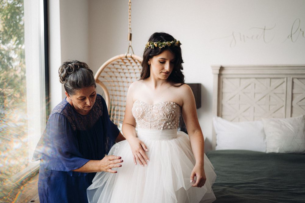 Intimate moment during wedding dress fitting in a sunlit room with rattan chair and textured white headboard.