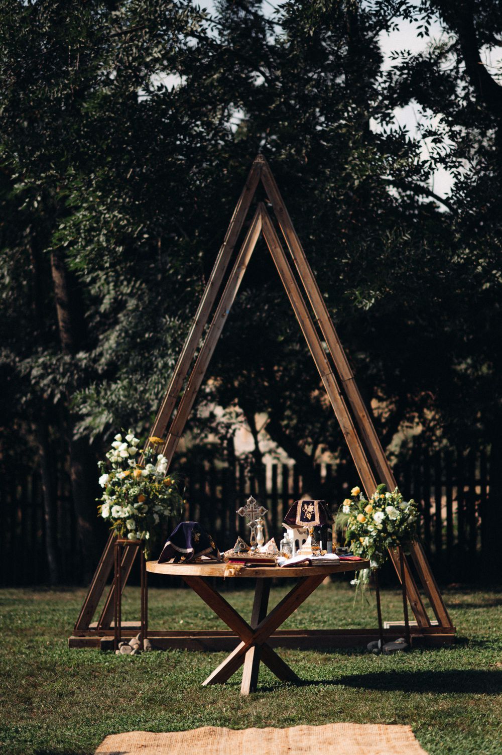 Wooden A-frame ceremony arch with floral arrangements and table display in outdoor forest setting.