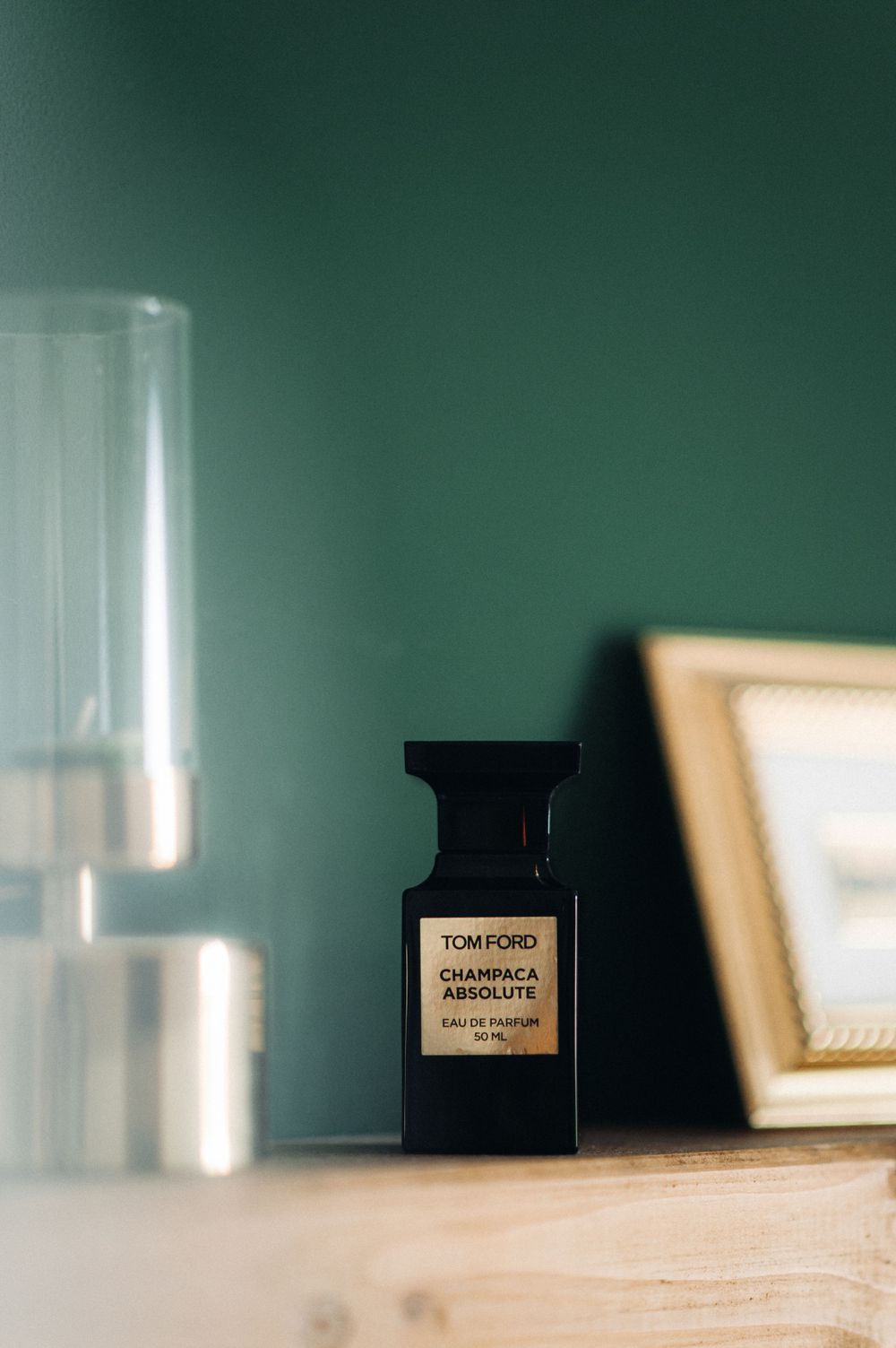 Tom Ford perfume bottle sits against a dark green wall with glass decor and frame in background.