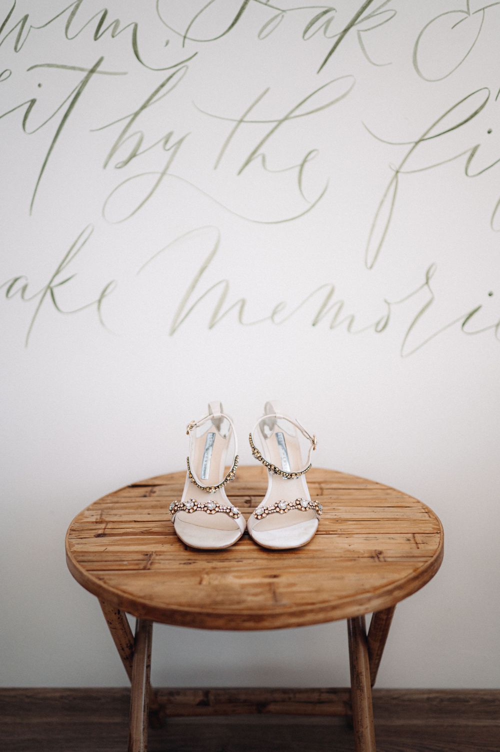 White bridal shoes with crystal details sit on a round wooden table beneath cursive wall text.