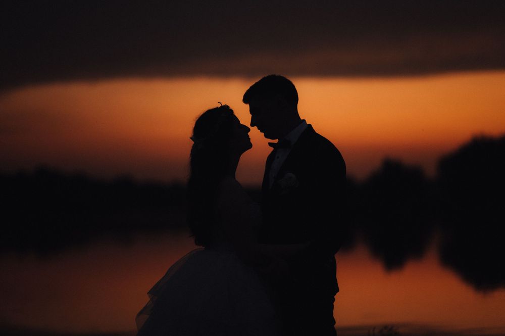 Silhouettes of a couple embracing against a dramatic orange sunset over water.