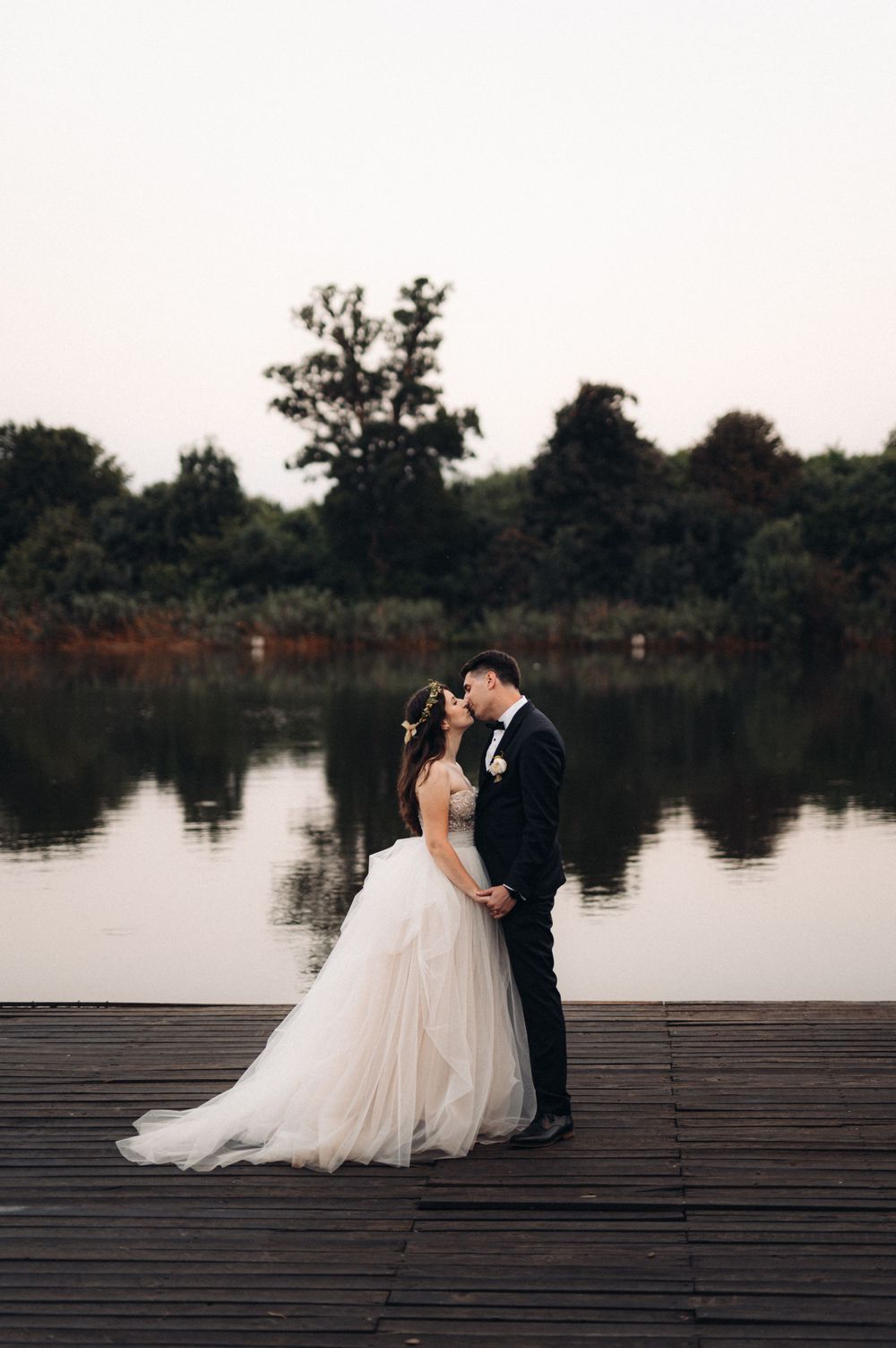 Wedding couple stands close together on wooden dock overlooking calm waters at dusk.