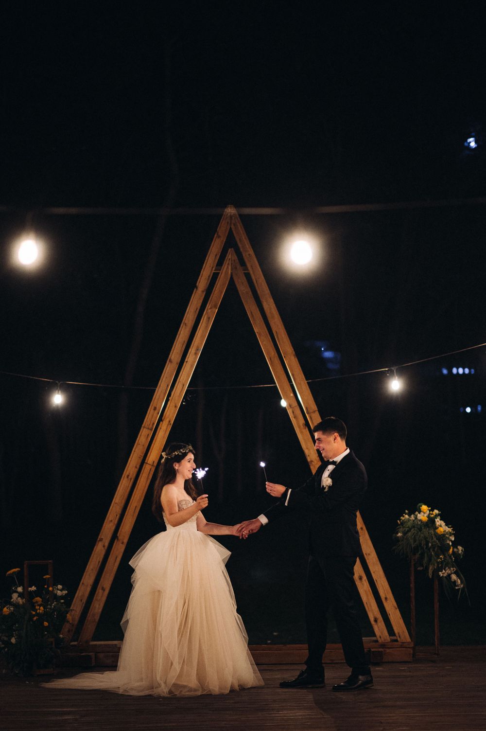 A couple dances under a wooden triangular arch with string lights at an evening wedding reception.
