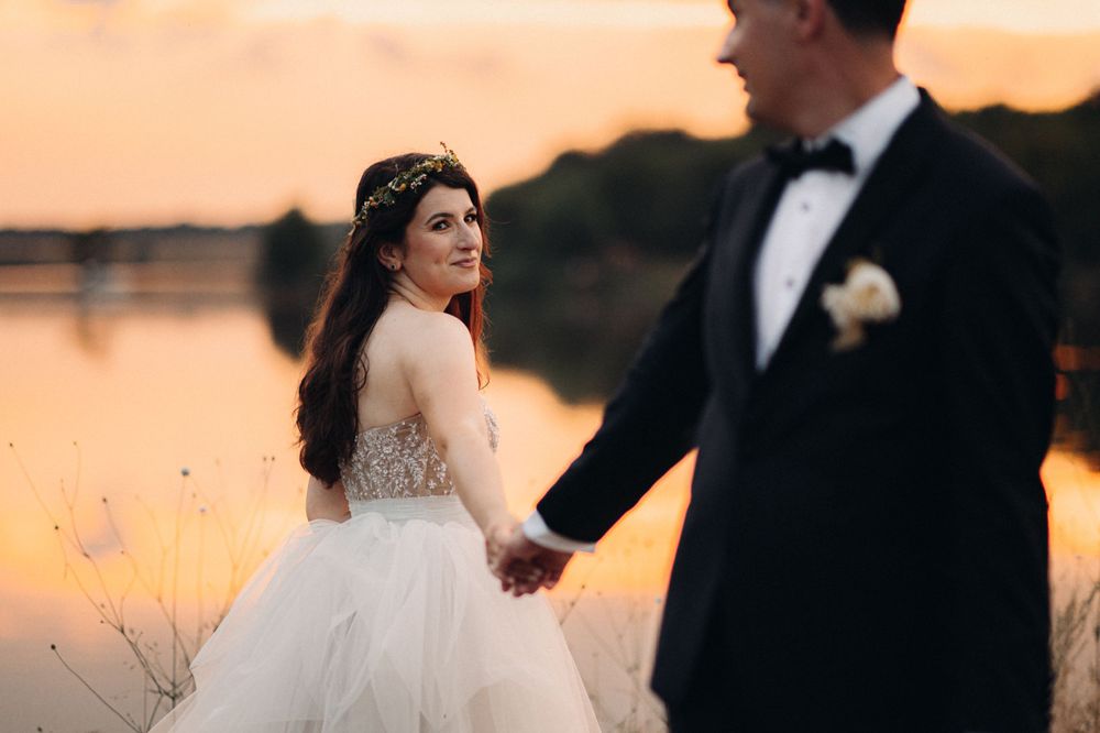 Bride in a flowing ballgown looks back while holding hands with groom during golden hour by the water.