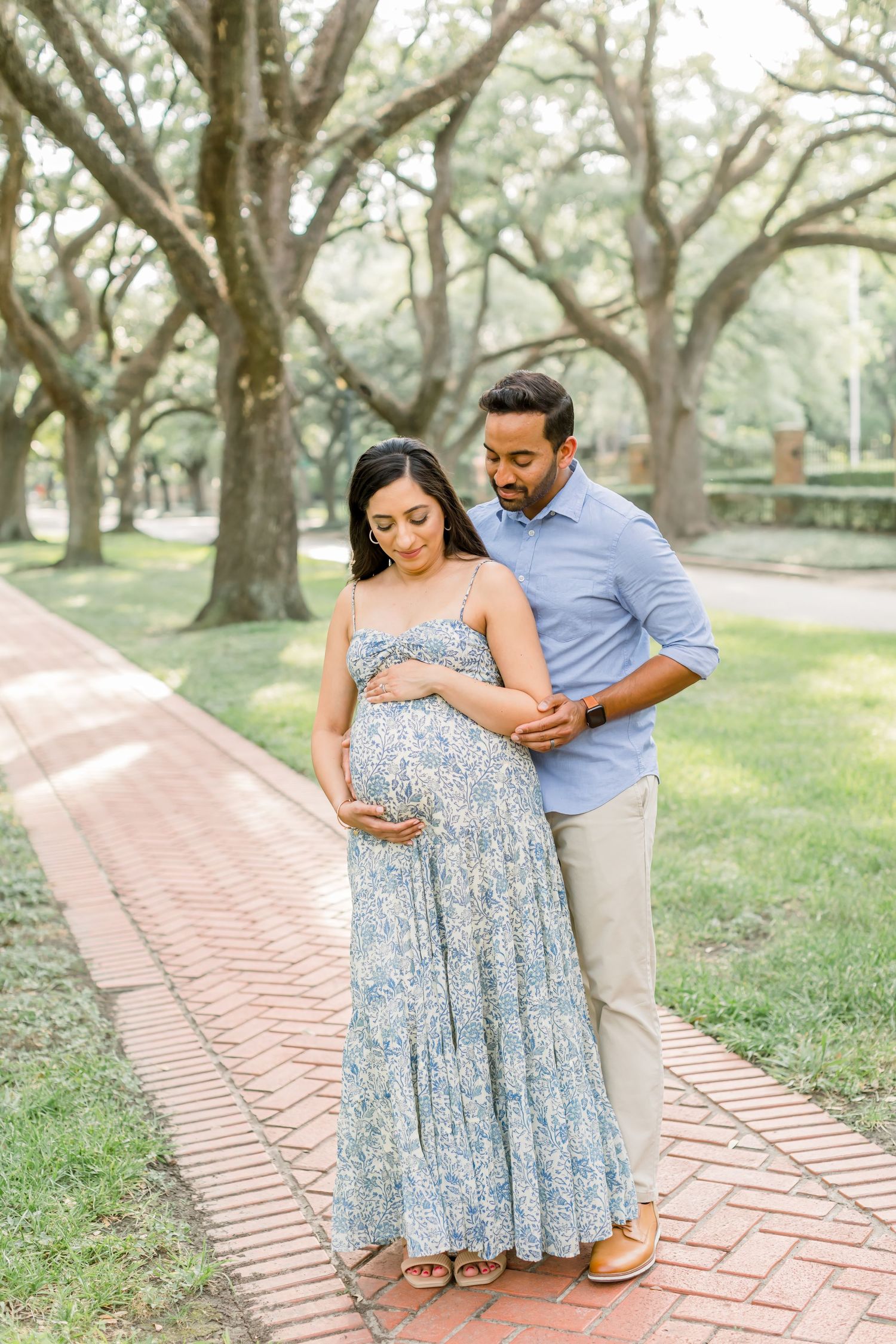 Intimate maternity portrait of a couple standing on red brick path beneath towering trees.