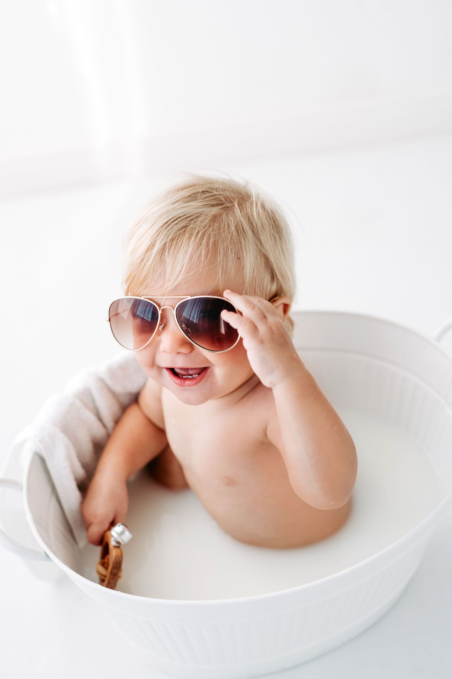 Joyful baby wearing oversized sunglasses sitting in white bowl.