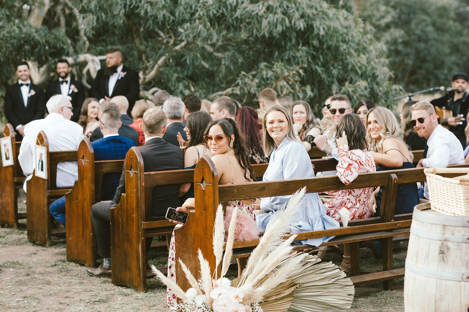 Wedding guests seated on wooden pews outdoors with dried pampas grass decorations.