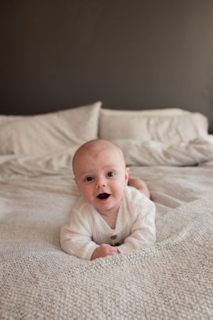 Smiling baby in white sweater lying on stomach on textured beige bedding.