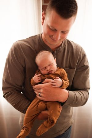A gentle embrace between parent and sleeping infant in brown clothing against soft window light.